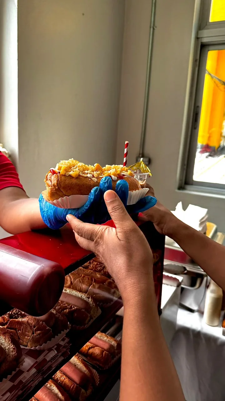 Entrega de un hot dog con papitas y salsas a un cliente junto al carrito rojo